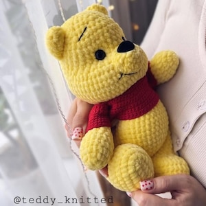 Op de afbeelding: Een gehaakt Winnie de Poeh knuffel met een rood shirt. Het speelgoed is geel en heeft een zwarte neus en ogen.