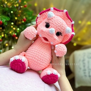May include: A pink crocheted triceratops stuffed animal with white horns and pink accents. The toy has black eyes and a pink mouth.