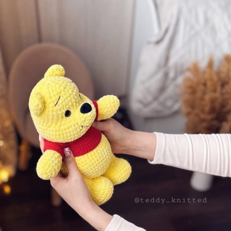 Op de afbeelding: Een gehaakt Winnie de Poeh knuffelbeest met een rood shirt. De beer is geel en heeft een bruine neus en zwarte ogen. De beer wordt vastgehouden door de handen van een persoon.