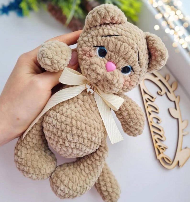 Puede incluir: Un oso de peluche hecho a mano, de color marr&oacute;n claro. El oso tiene ojos azules, una nariz rosa y una cinta color crema alrededor del cuello. El oso est&aacute; siendo sostenido.
