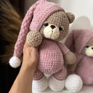 以下が含まれることがあります： ピンクの編みぐるみ帽子とパジャマを着た、編みぐるみのテディベア。クマは茶色の毛皮で、顔に白いアクセントがあります。クマは左の足に白い編みぐるみのボールを持っています。
