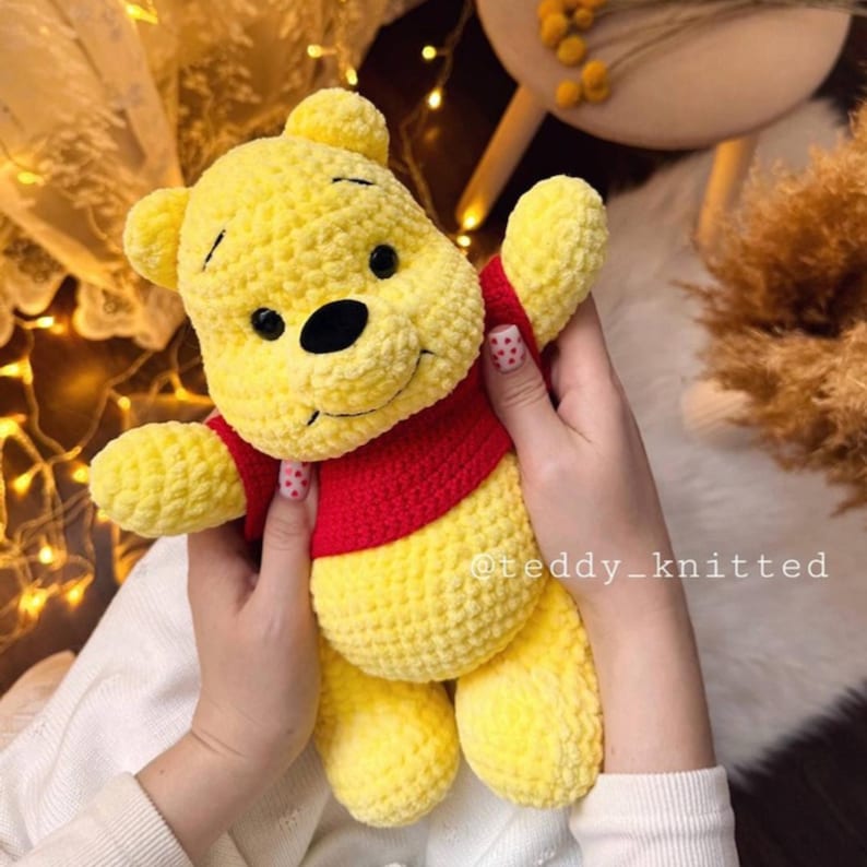 May include: A crocheted Winnie the Pooh plush toy with a yellow body, red shirt, and black eyes. The toy is being held by a person's hands.