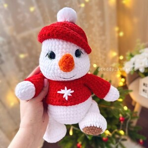 May include: A crocheted snowman toy wearing a red hat and a red sweater with a white snowflake design. The snowman has a white body, orange nose, and black eyes.