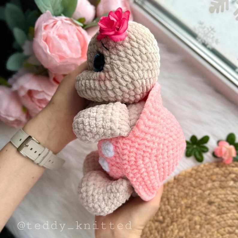 May include: A crocheted plush toy in the shape of a turtle wearing a pink crocheted dress with white polka dots. The turtle has a pink flower on its head and large blue eyes.