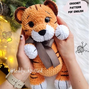 May include: A crocheted tiger toy in orange and brown, with a white face, paws, and belly. It has a brown bow and black eyes. The image includes the text "CROCHET PATTERN PDF ENGLISH" in a cloud-shaped graphic.