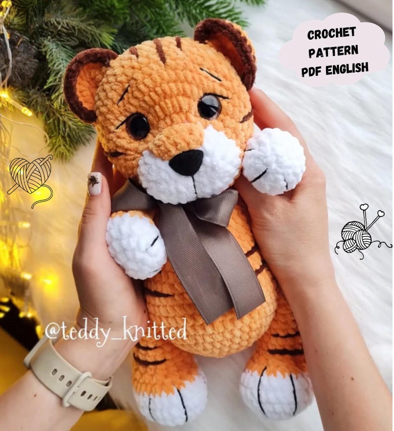 May include: A handmade orange and brown crochet tiger plushie with a white muzzle, paws, and belly. The tiger has a brown bow and black eyes. The text "CROCHET PATTERN PDF ENGLISH" is in a cloud-shaped graphic.