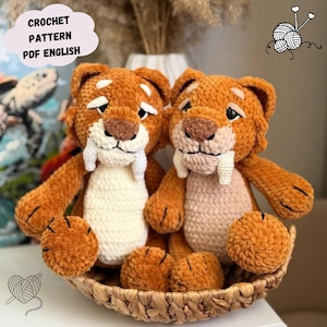 Könnte beinhalten: Zwei handgefertigte Häkeltier-Plüschtiere eines Säbelzahntigers in einem Weidenkorb. Die Tiger sind orange mit weißen Stoßzähnen und cremefarbenen Bäuchen. Der Text "CROCHET PATTERN PDF ENGLISH" befindet sich in einer Wolkenblase.