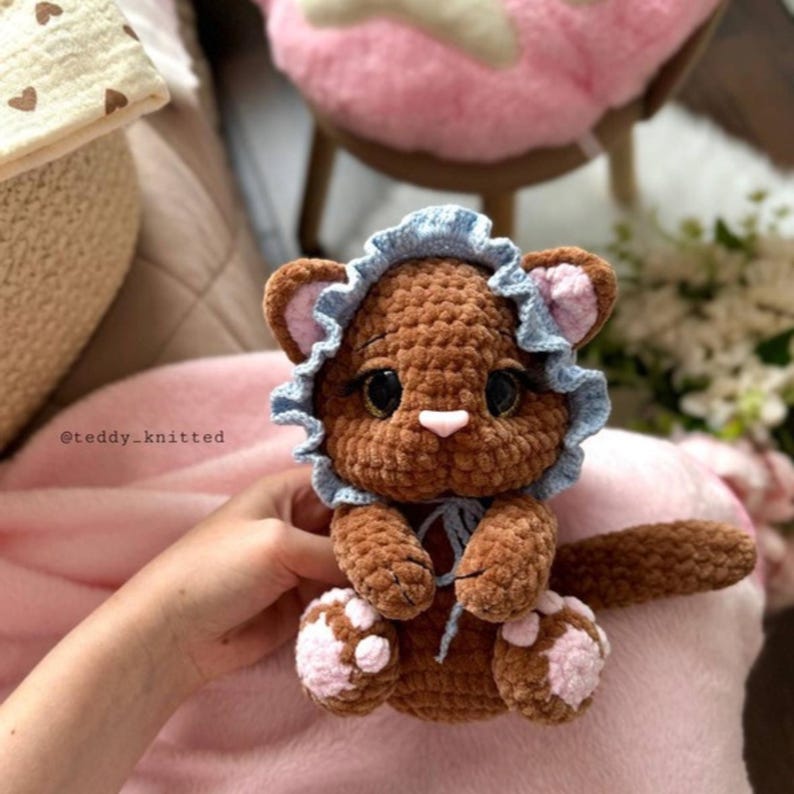 May include: A handmade brown crochet lion cub wearing a blue bonnet with a ruffled edge. The lion has pink accents on its ears and paws, and large black eyes. The text "@teddy_knitted" is visible.