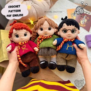 Pode incluir: Três bonecos de crochê feitos à mão com cabelo de lã e roupas. O boneco da esquerda veste um suéter vermelho com um R, o do meio um suéter verde com saia cinza e o da direita um suéter azul com um H. Todos usam cachecóis vermelhos e amarelos.