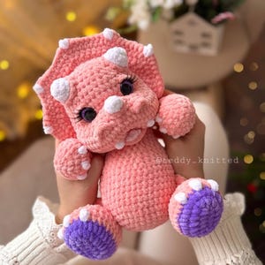 May include: A pink crocheted triceratops stuffed animal with purple and white feet. The triceratops has white horns and black eyes.