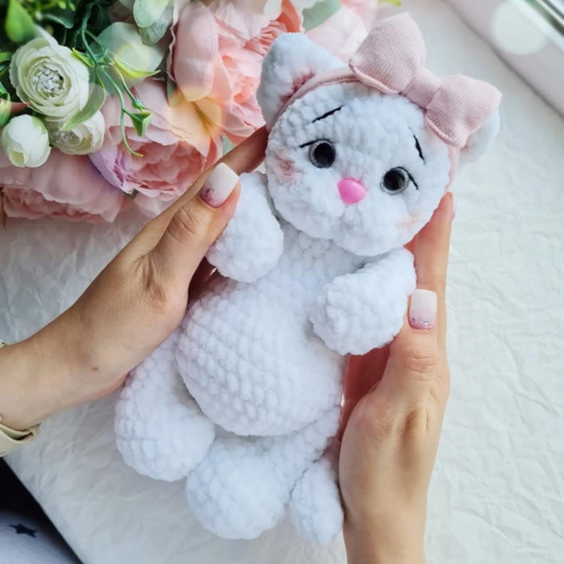 Puede incluir: Una mu&ntilde;eca de gato de ganchillo blanco hecha a mano con un lazo y una diadema rosas. El gato tiene ojos negros, nariz rosa y colorete. La mu&ntilde;eca se sostiene con dos manos, con un fondo floral.