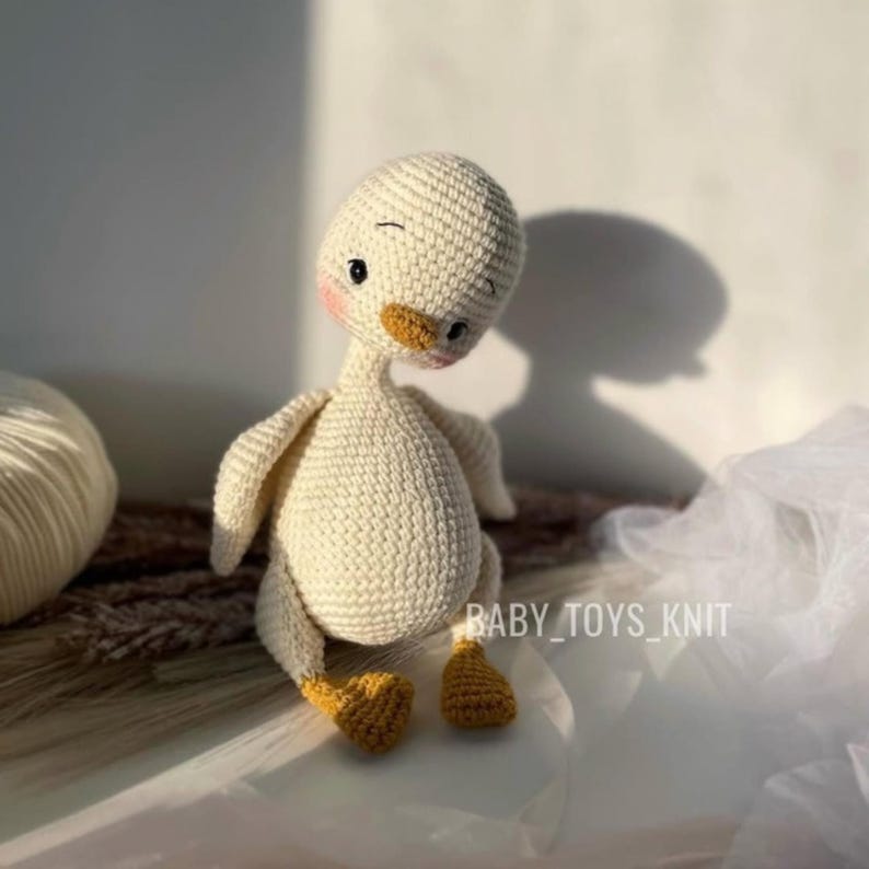 May include: A crocheted white duck with yellow feet and a small orange beak. The duck has a pink blush on its cheeks and black eyes. The duck is sitting on a light brown surface with a white fabric in the background. The text "BABY_TOYS_KNIT" is visible in the bottom right corner of the image.