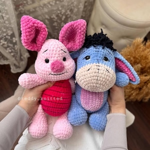 May include: Two crocheted stuffed animals, a pink pig and a blue donkey, are being held by a person's hands. The pig has a red and white striped shirt. The donkey has black hair on its head. The stuffed animals are made with a soft, fuzzy yarn.