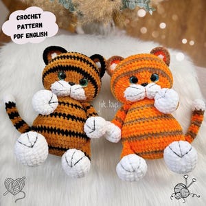 Pode incluir: Dois brinquedos de pelúcia de tigre de crochê. Os tigres são laranja com listras pretas, patas brancas e olhos verdes ou azuis. O texto "CROCHET PATTERN PDF ENGLISH" está em uma bolha em forma de nuvem. Os brinquedos são feitos à mão.