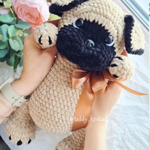 May include: A crocheted plush pug toy with black and beige fur, brown eyes, and a brown ribbon around its neck. The toy is being held by a person's hands. The text "@teddy_knitted" is visible on the ribbon.