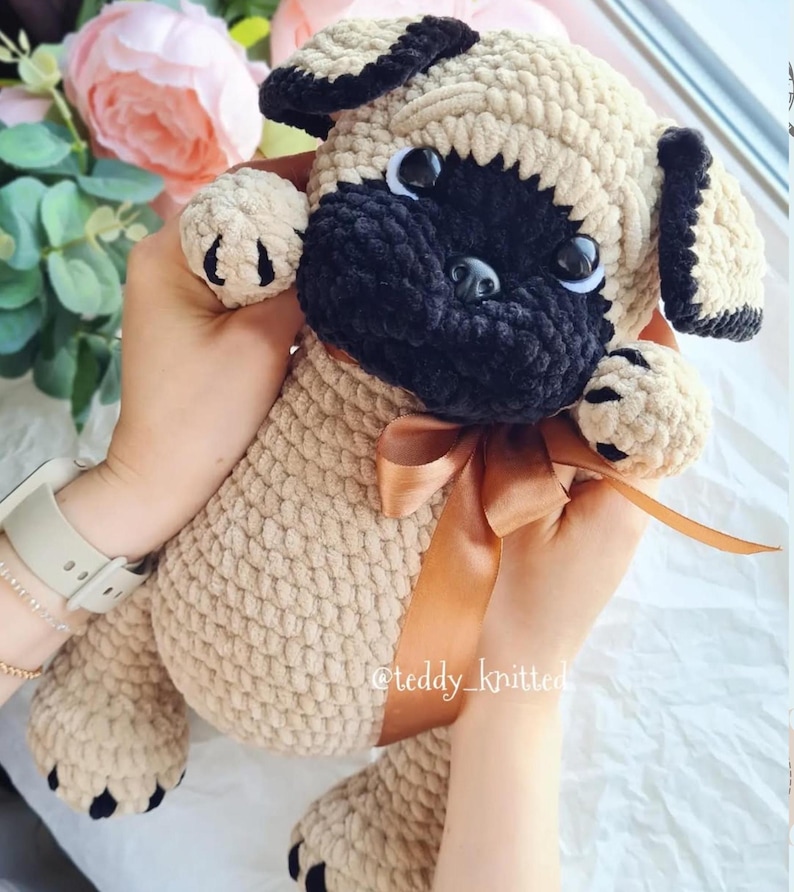 May include: A crocheted plush pug toy with black and beige fur, brown eyes, and a brown ribbon around its neck. The toy is being held by a person's hands. The text "@teddy_knitted" is visible on the ribbon.