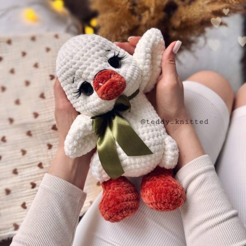 May include: A handmade white crochet duck plushie with a red beak and feet, black eyelashes, and a green ribbon. The plushie is held in someone's hands. The text "@teddy_knitted" is visible.