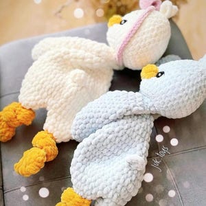 Op de afbeelding: Twee gehaakte knuffels in de vorm van vogels. E&eacute;n is wit met een roze strik en de andere is lichtblauw. Beide hebben gele voeten.