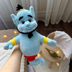 May include: A light blue crocheted plush toy shaped like a genie with a black beard, black eyebrows, and a black topknot. The figure features yellow wrist cuffs, a red waist section, and a curved lower body. The text @teddy_knitted is visible.