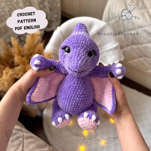May include: A handmade purple crochet pterodactyl plush toy with pink wings and accents. The toy has large black eyes and a smiling expression. Text on the image reads "CROCHET PATTERN PDF ENGLISH". The toy is held in front of a soft, blurred background.