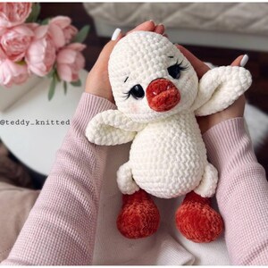 May include: A handmade white crochet duck with a red beak and feet. The duck has black eyes with long eyelashes and is being held by a person wearing a pink sweater. The duck's wings are outstretched.