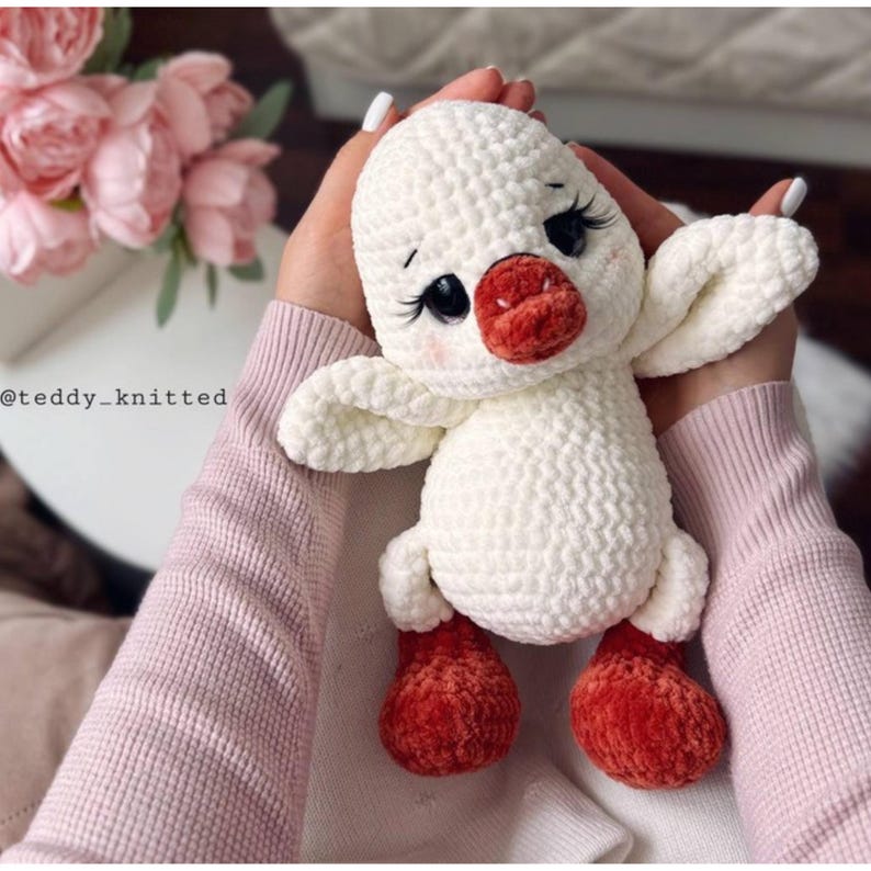 May include: A handmade white crochet duck with a red beak and feet. The duck has black eyes with long eyelashes and is being held by a person wearing a pink sweater. The duck's wings are outstretched.