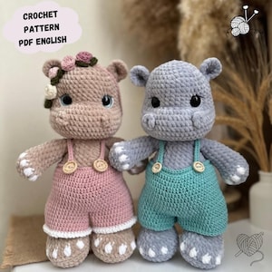 Op de afbeelding: Twee gehaakte nijlpaard speelgoedjes die overalls dragen. Een nijlpaard is roze met een bloemenhoofdband en de andere is grijs met blauwe overalls.