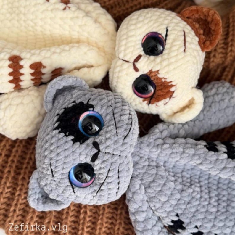May include: Three handmade crochet teddy bears in cream and gray. The bears have large, iridescent eyes and stitched details. One bear has brown patches and ears. The bears are lying on a brown surface.