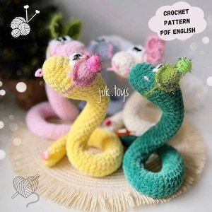 Puede incluir: Juguetes de serpiente tejidos a ganchillo en amarillo y verde, con detalles en rosa y verde. La imagen incluye el texto "CROCHET PATTERN PDF ENGLISH" y el nombre de la marca "juk.toys". Estos juguetes son hechos a mano.