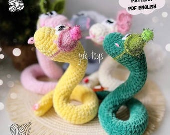 May include: Crocheted snake toys in yellow and green, with pink and green accents. The toys have a whimsical design, with the text "CROCHET PATTERN PDF ENGLISH" in a cloud-shaped graphic. The brand name "juk.toys" is also visible.