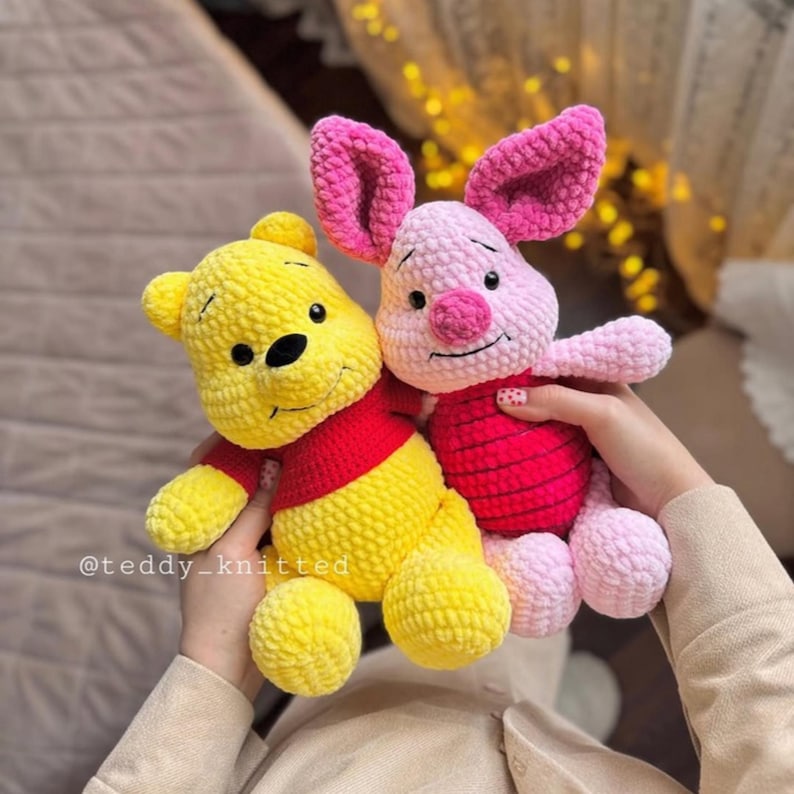 May include: A yellow crocheted Winnie the Pooh plush toy and a pink crocheted Piglet plush toy. Both toys have embroidered facial features and are holding hands.