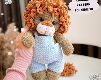 May include: Crocheted lion amigurumi toy wearing blue overalls. The lion has a fluffy orange mane and tail. The image includes the text "CROCHET PATTERN PDF ENGLISH".