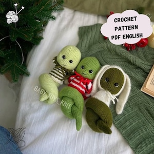 Pode incluir: Bonecas alienígenas de crochê feitas à mão em tons de verde, branco e vermelho. Uma boneca tem aparência de coelho. As bonecas estão deitadas em uma superfície branca com um cobertor de malha verde. O texto na imagem diz "CROCHET PATTERN PDF ENGLISH" e "BABY TOYS KNIT".