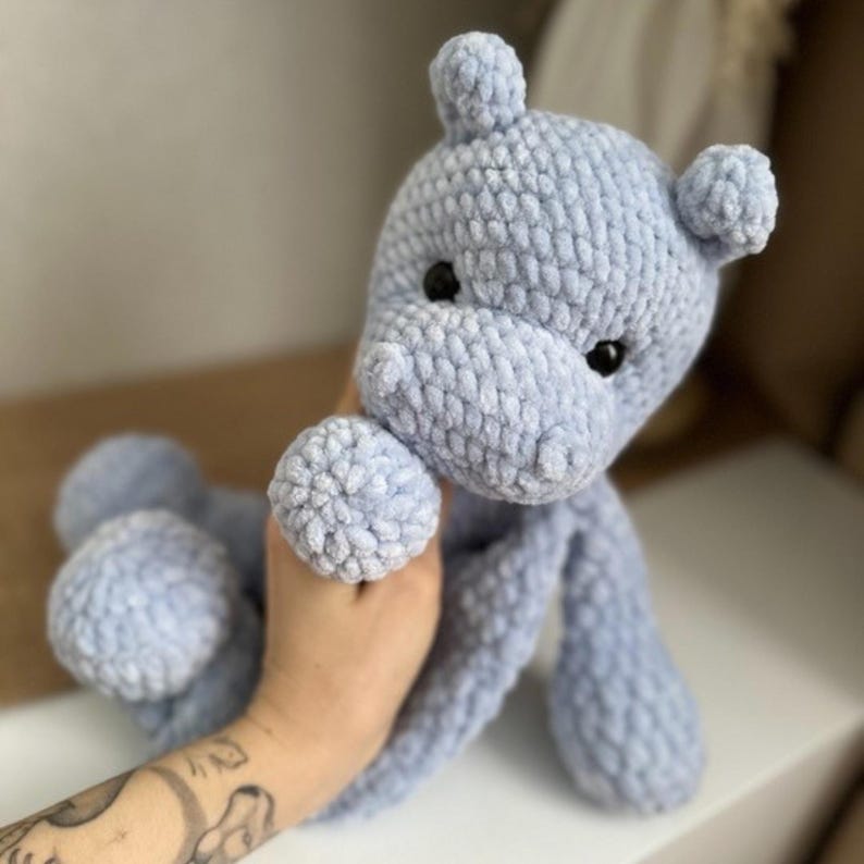 Puede incluir: Un juguete de peluche de hipop&oacute;tamo de color azul claro tejido a crochet. El hipop&oacute;tamo tiene una superficie texturizada y peluda y ojos negros.