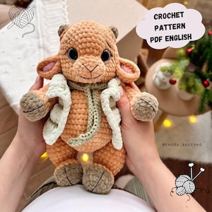 Puede incluir: Un peluche de cordero tejido a ganchillo en tonos beige, marrón y crema. Tiene una bufanda verde y una chaqueta color crema. El texto "CROCHET PATTERN PDF ENGLISH" está en un bocadillo. El juguete está en manos.