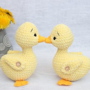 Peut inclure: Deux canetons au crochet jaunes faits &agrave; la main avec des becs et des pieds orange se touchent le bec. Les canetons ont des yeux noirs et de petits boutons en bois. Le fond est gris clair.