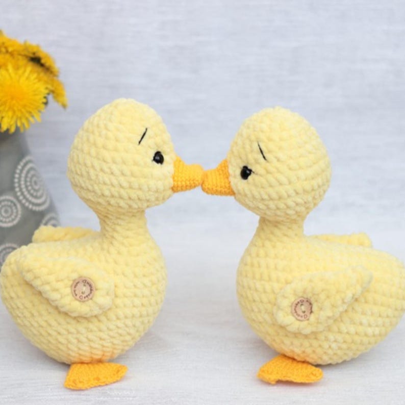 Peut inclure: Deux canetons au crochet jaunes faits &agrave; la main avec des becs et des pieds orange se touchent le bec. Les canetons ont des yeux noirs et de petits boutons en bois. Le fond est gris clair.