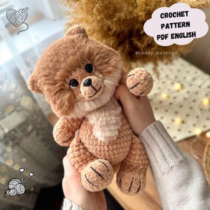 Puede incluir: Un oso de peluche de ganchillo en tonos marrón y beige, con ojos negros y una nariz cosida. El oso se sostiene en las manos y presenta el texto "CROCHET PATTERN PDF ENGLISH" en una nube.