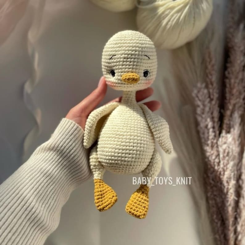 May include: A white crocheted duck toy with yellow feet and a black embroidered face. The duck is being held by a hand.