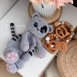 May include: A gray plush cat toy with black markings and pink paw pads. Next to the cat is a wooden baby rattle with a cat design and wooden beads. The text "Zefirka.vlg" is visible.