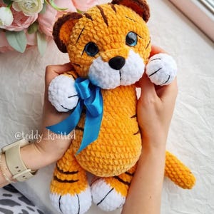 May include: A handmade orange and brown tiger plush toy with a blue ribbon. The tiger has black stripes, white paws, and blue eyes. The toy is held by a person. The text "@teddy_knitted" is visible on the left wrist.