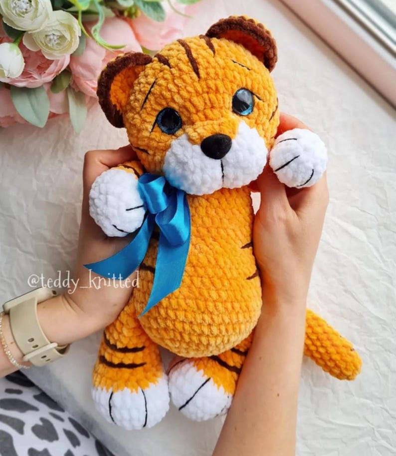 May include: A handmade orange and brown tiger plush toy with a blue ribbon. The tiger has black stripes, white paws, and blue eyes. The toy is held by a person. The text "@teddy_knitted" is visible on the left wrist.