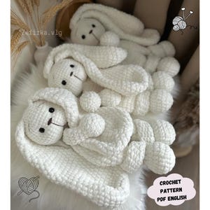 May include: Three white crocheted bunny amigurumi toys with long ears. The bunnies are lying on a white fluffy surface. The text "CROCHET PATTERN PDF ENGLISH" is visible in the bottom right corner of the image.