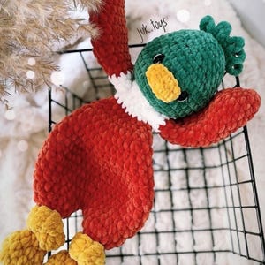 Op de afbeelding: Een gehaakt pluche speelgoed in de vorm van een eend. De eend is rood, groen, wit en geel. De eend ligt op zijn rug in een zwarte draadmand. De tekst "juk.toys" is zichtbaar op de eend.