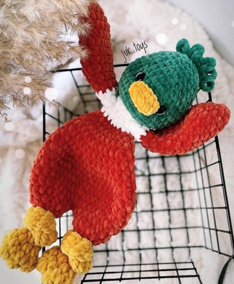 Op de afbeelding: Een gehaakt pluche speelgoed in de vorm van een eend. De eend is rood, groen, wit en geel. De eend ligt op zijn rug in een zwarte draadmand. De tekst "juk.toys" is zichtbaar op de eend.