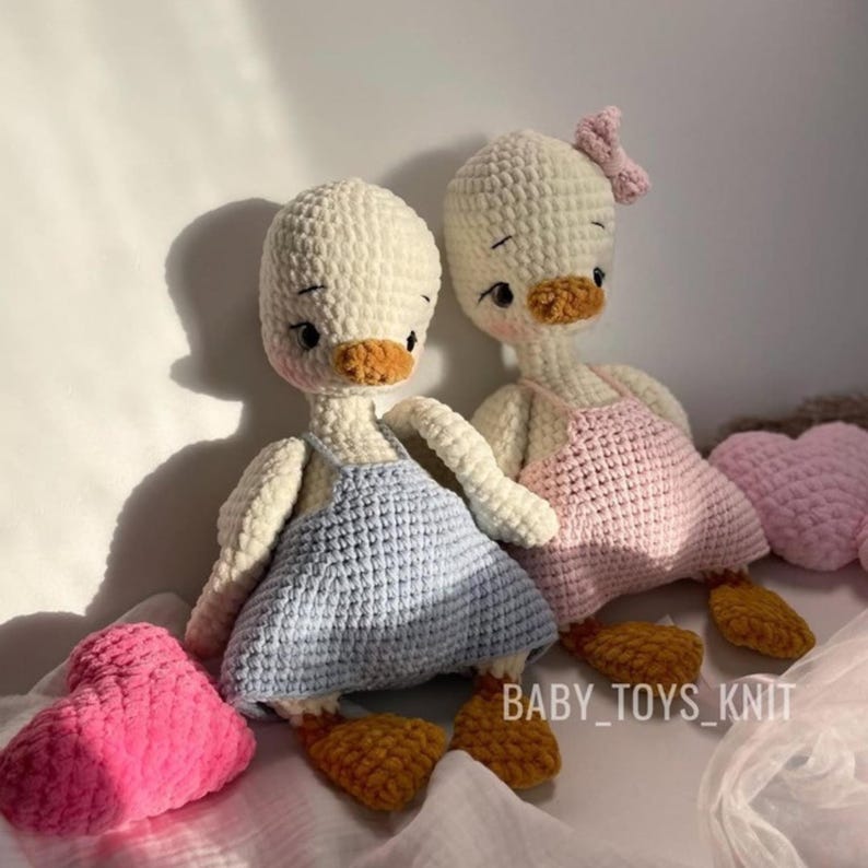 May include: Two crocheted duck plushies, one in white with a blue dress and the other in white with a pink dress. Both ducks have brown beaks and feet. The ducks are sitting on a pink and white fabric background.