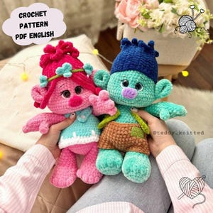 May include: Two handmade crochet troll dolls. One doll is pink with a blue dress and the other is teal with brown shorts. The dolls have large ears, colorful hair, and are held in front of a soft background. Text in the image reads "CROCHET PATTERN PDF ENGLISH".