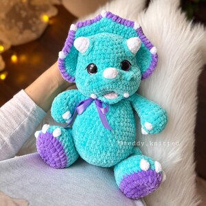 May include: A teal blue crocheted triceratops stuffed animal with purple and white accents. The dinosaur has a purple ribbon around its neck and white spots on its body.
