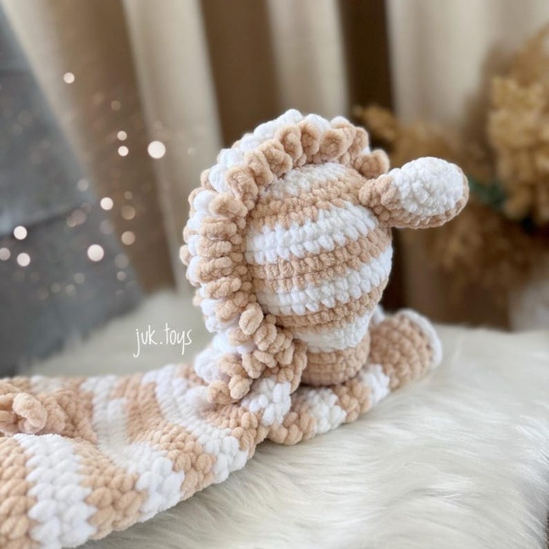May include: A handmade, plush toy in a light brown and white striped pattern. The toy has a textured, soft appearance and features a detailed mane and head. The text "juk.toys" is visible on the toy.