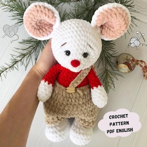 May include: Crocheted white mouse plush toy wearing a red shirt and brown overalls. The toy has a red nose and black eyes. The image includes the text "CROCHET PATTERN PDF ENGLISH".